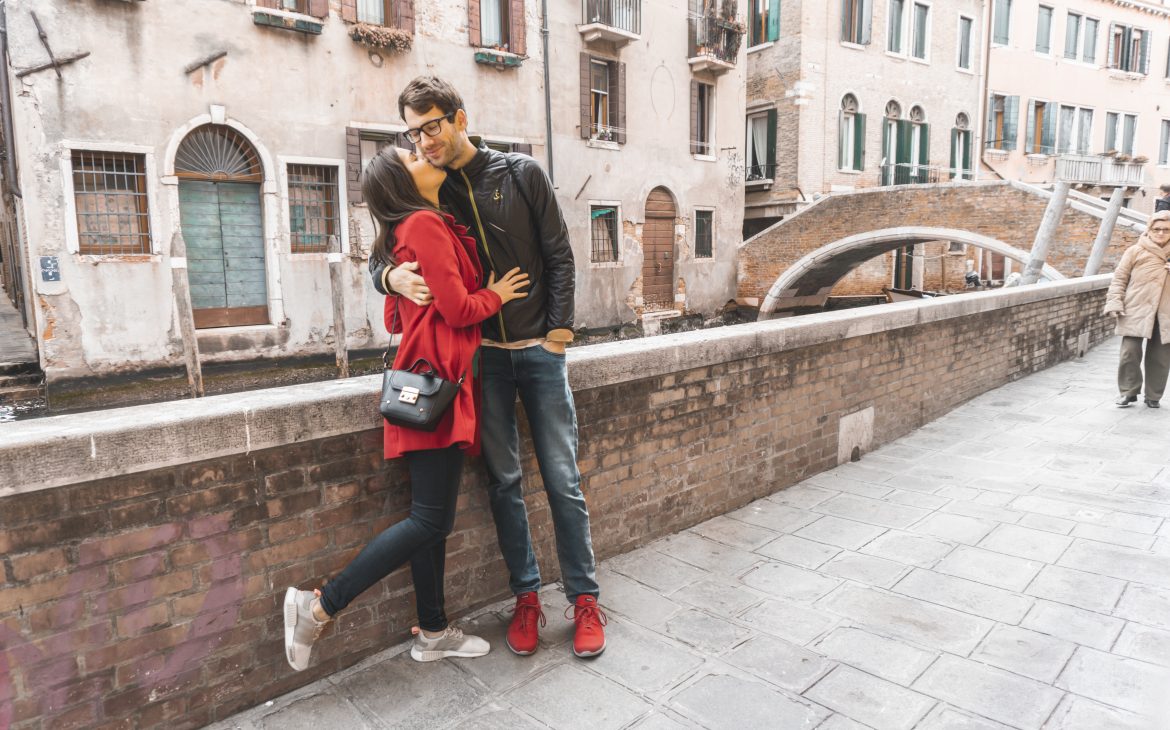 A girl in the red coat kissing the boy in the cheek in Venice. 