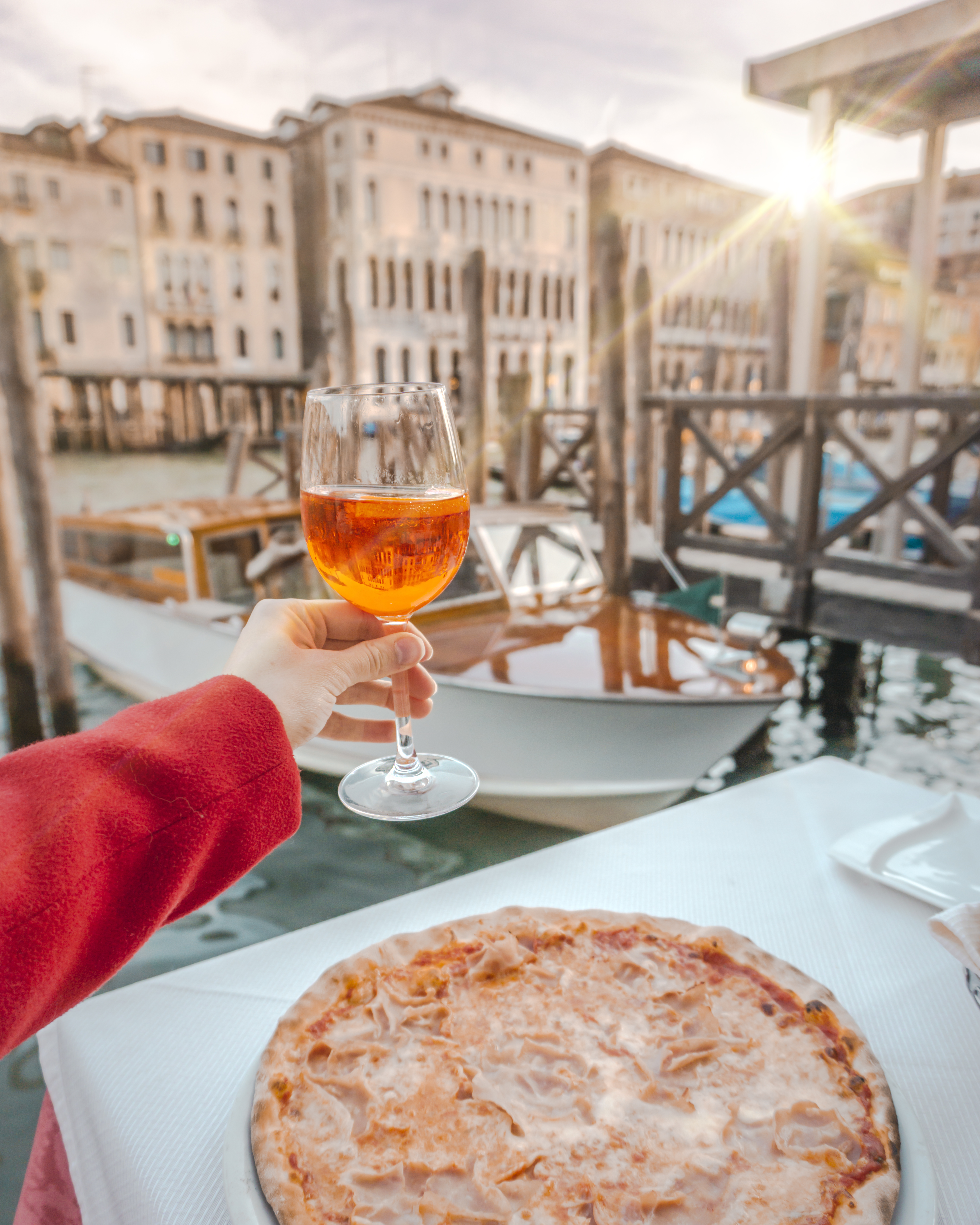 A person holding an orange Aperol drink in Venice with the pizza on the table. 