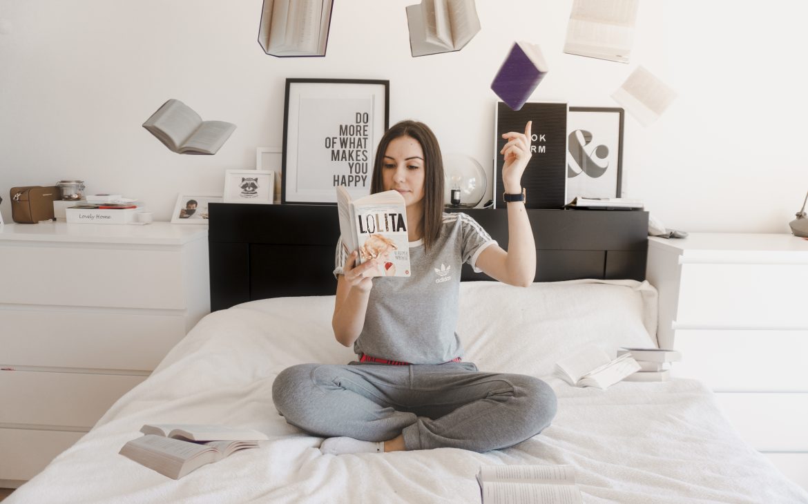 a girl sitting on the bed and reading book, while having flying books above her head